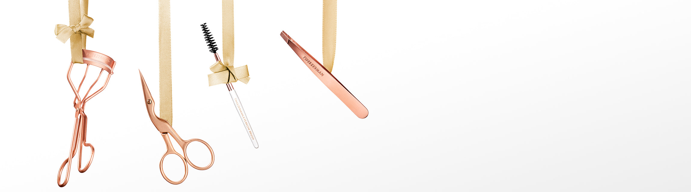 A set of rose gold beauty tools hanging from gold ribbon against a white background, including an eyelash curler, brow scissors, a brow spoolie, and a mini slant tweezer