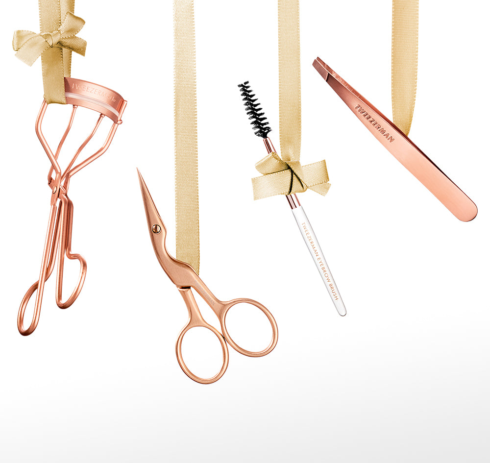 A set of rose gold beauty tools hanging from gold ribbon against a white background, including an eyelash curler, brow scissors, a brow spoolie, and a mini slant tweezer