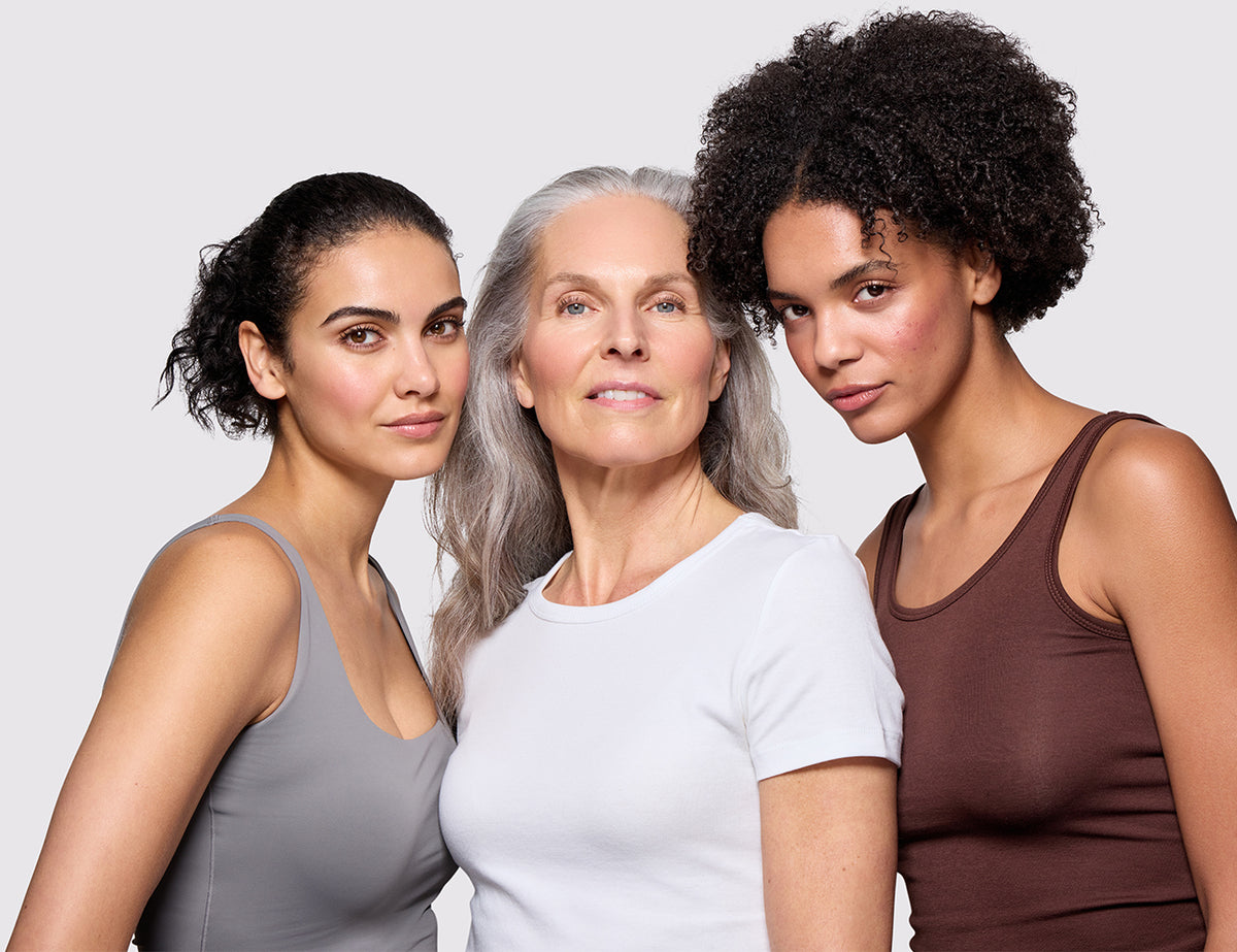 Three women standing together against a light gray studio background, wearing sleeveless tank tops in gray, white, and brown.