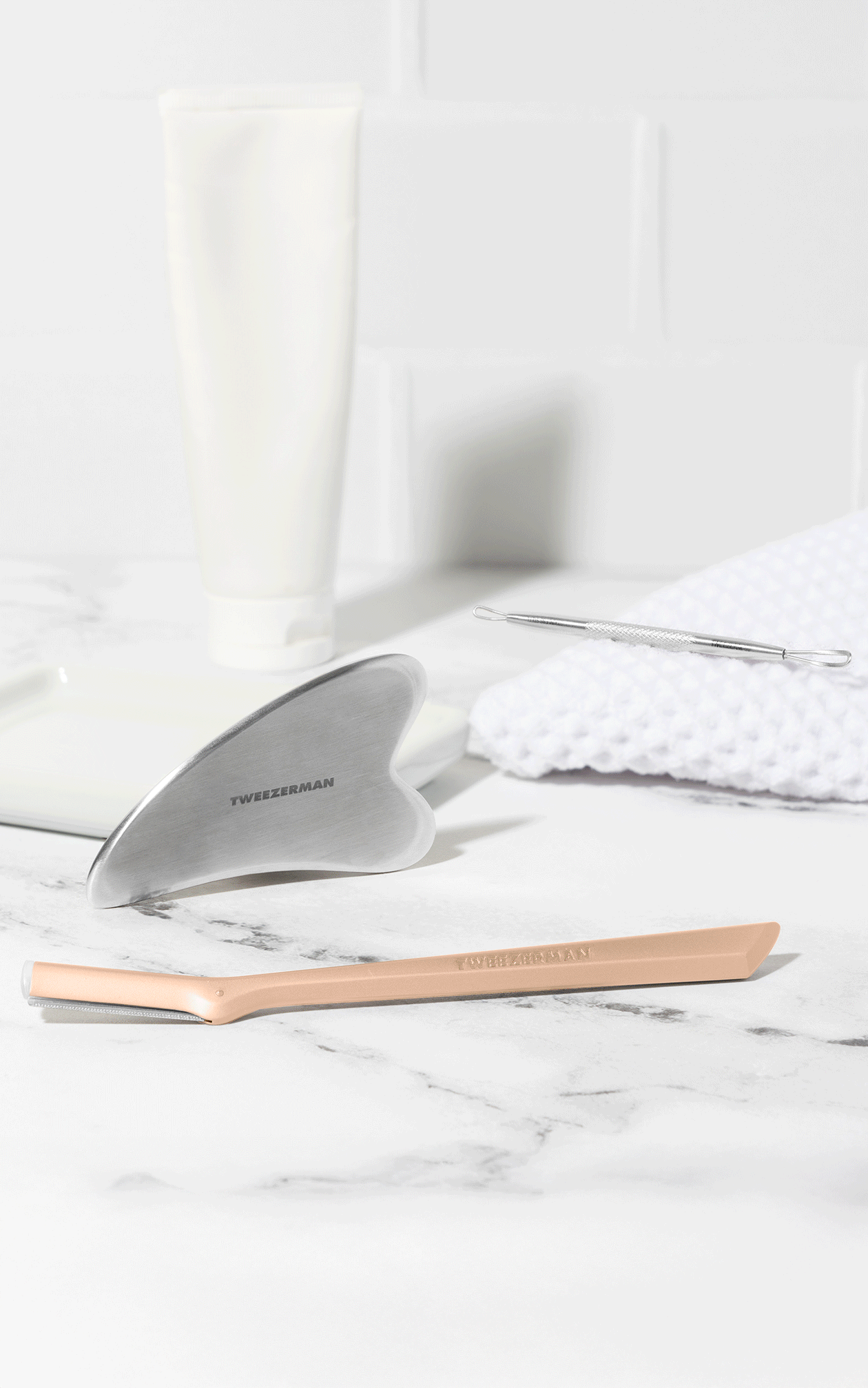 Stainless Steel Gua Sha, Neon Nail Files and combo clipper set, Stainless steel wide grip lash applicator, mini slant tweezer and brow razor. Rose gold facial razor. background is a vanity with towel and cuticle oil.