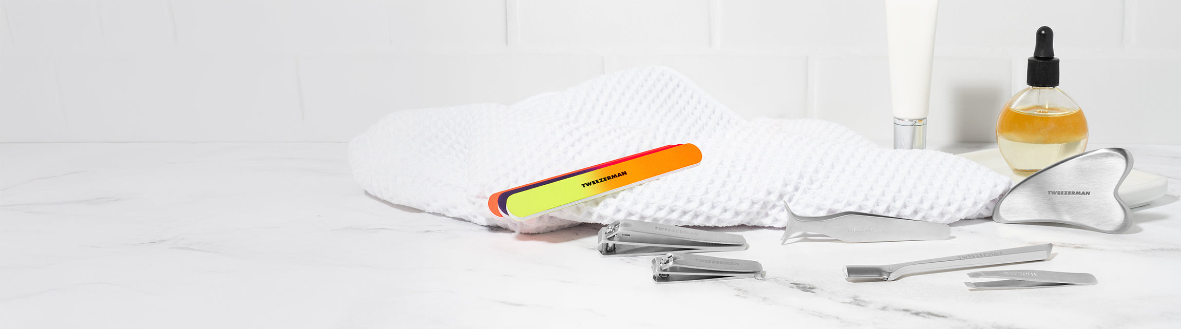 Stainless Steel Gua Sha, Neon Nail Files and combo clipper set, Stainless steel wide grip lash applicator, mini slant tweezer and brow razor. background is a vanity with towel and cuticle oil.