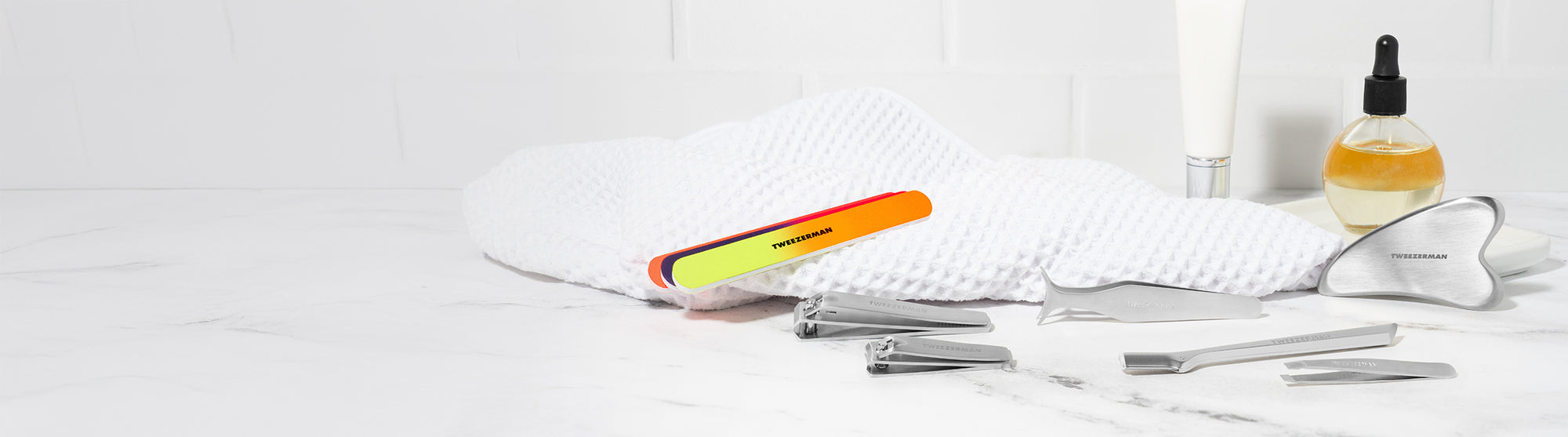 Stainless Steel Gua Sha, Neon Nail Files and combo clipper set, Stainless steel wide grip lash applicator, mini slant tweezer and brow razor. background is a vanity with towel and cuticle oil.