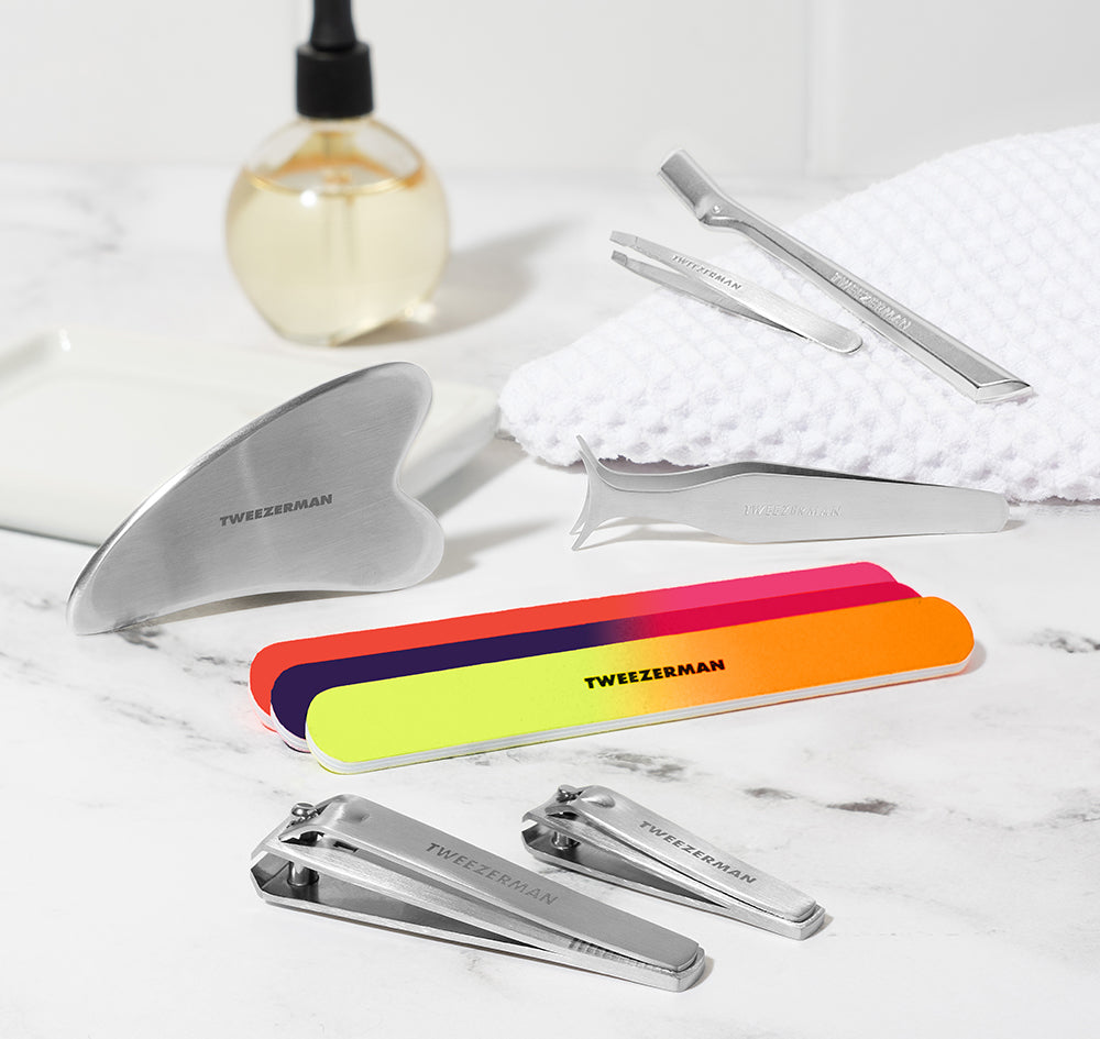 Stainless Steel Gua Sha, Neon Nail Files and combo clipper set, Stainless steel wide grip lash applicator, mini slant tweezer and brow razor. background is a vanity with towel and cuticle oil.